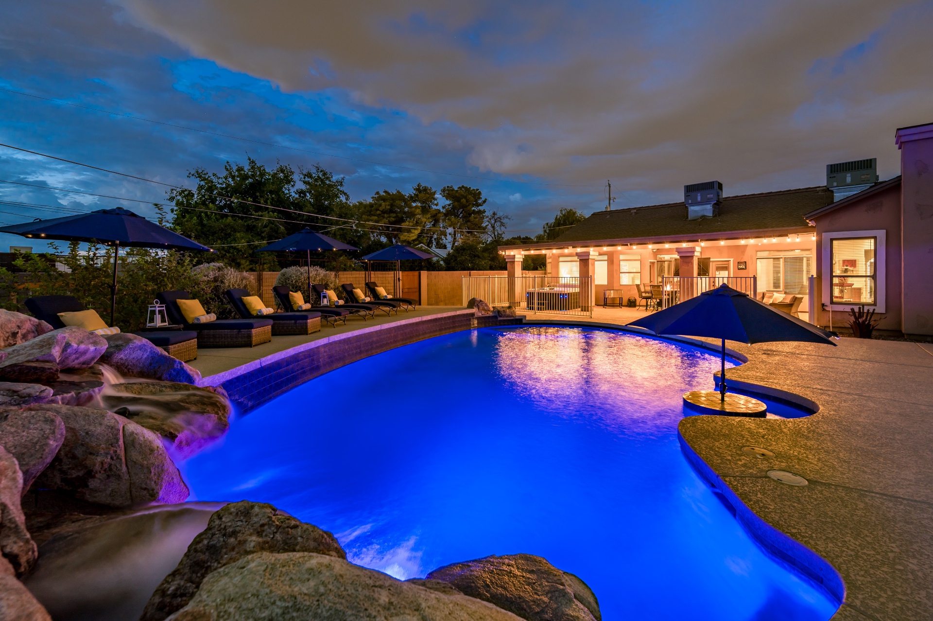Pool glowing blue at dusk with loungers, umbrellas and string-lit patio
